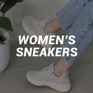 Een paar witte sneakers die gedragen worden door een mevrouw, ze zit in een witte kamer met een plant in de hoek.