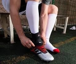 Sporter op een sportveld trekt een enkelbrace aan over sportsokken, terwijl hij voetbalschoenen draagt