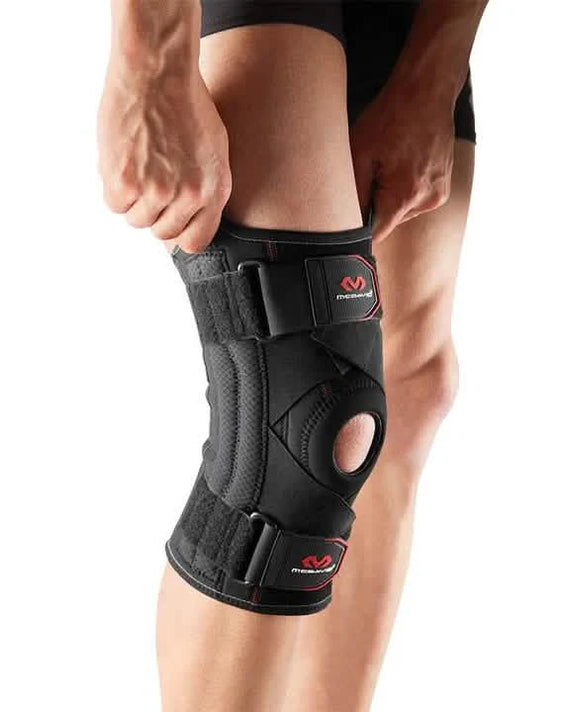 McDavid-425-Knee-Support-With-Stays-&-Cross-Straps-Person adjusting a black knee brace with straps on their right knee-The brace has a circular opening at the kneecap.