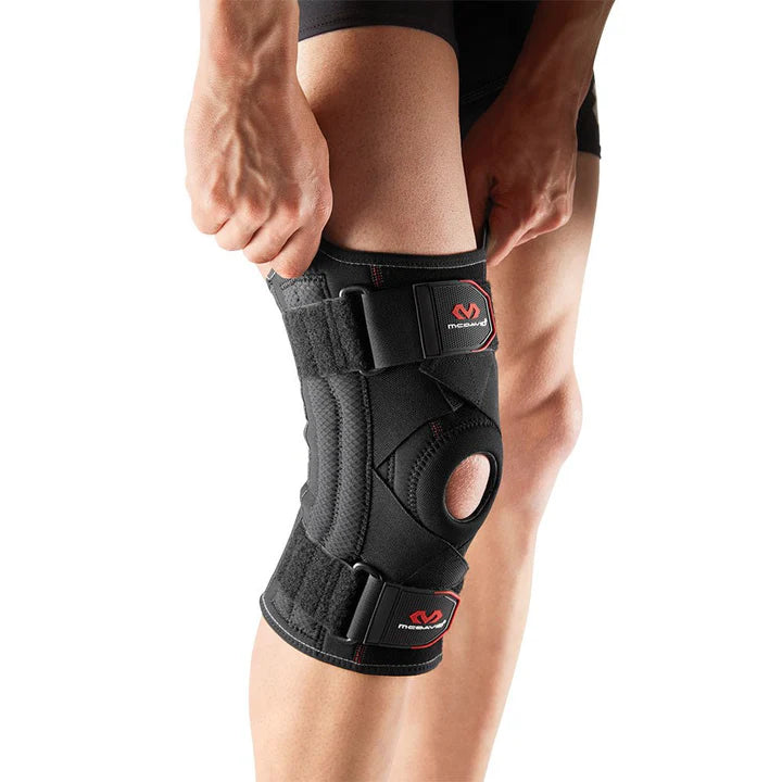 McDavid-425-Knee-Support-With-Stays-&-Cross-Straps-Person adjusting a black knee brace with straps on their right knee-The brace has a circular opening at the kneecap.
